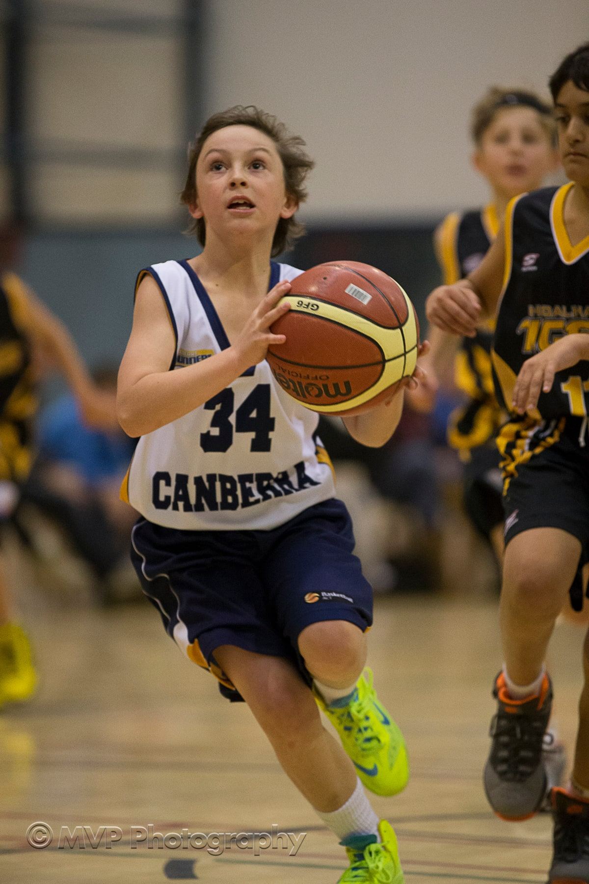 A young player in a Canberra representative jersey catches a pass on court, closely followed by defenders.