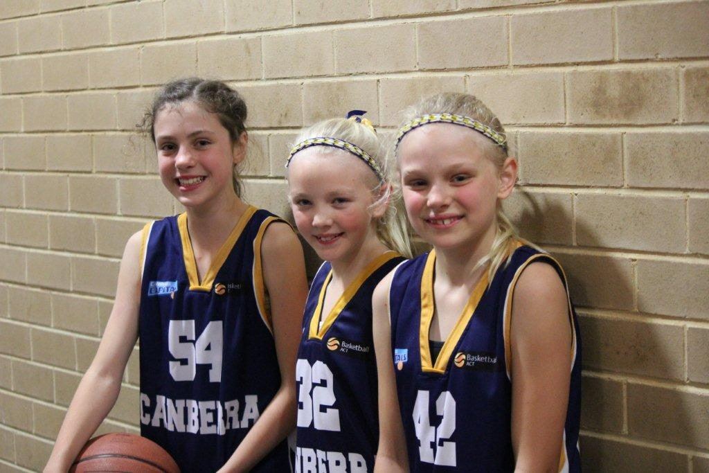Three smiling girls in navy Canberra representative jerseys stand together against a brick wall, one holding a basketball.