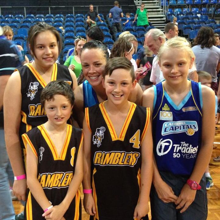 Ramblers junior players and a Canberra Capitals fan pose together with an adult at a stadium, bleachers in the background.