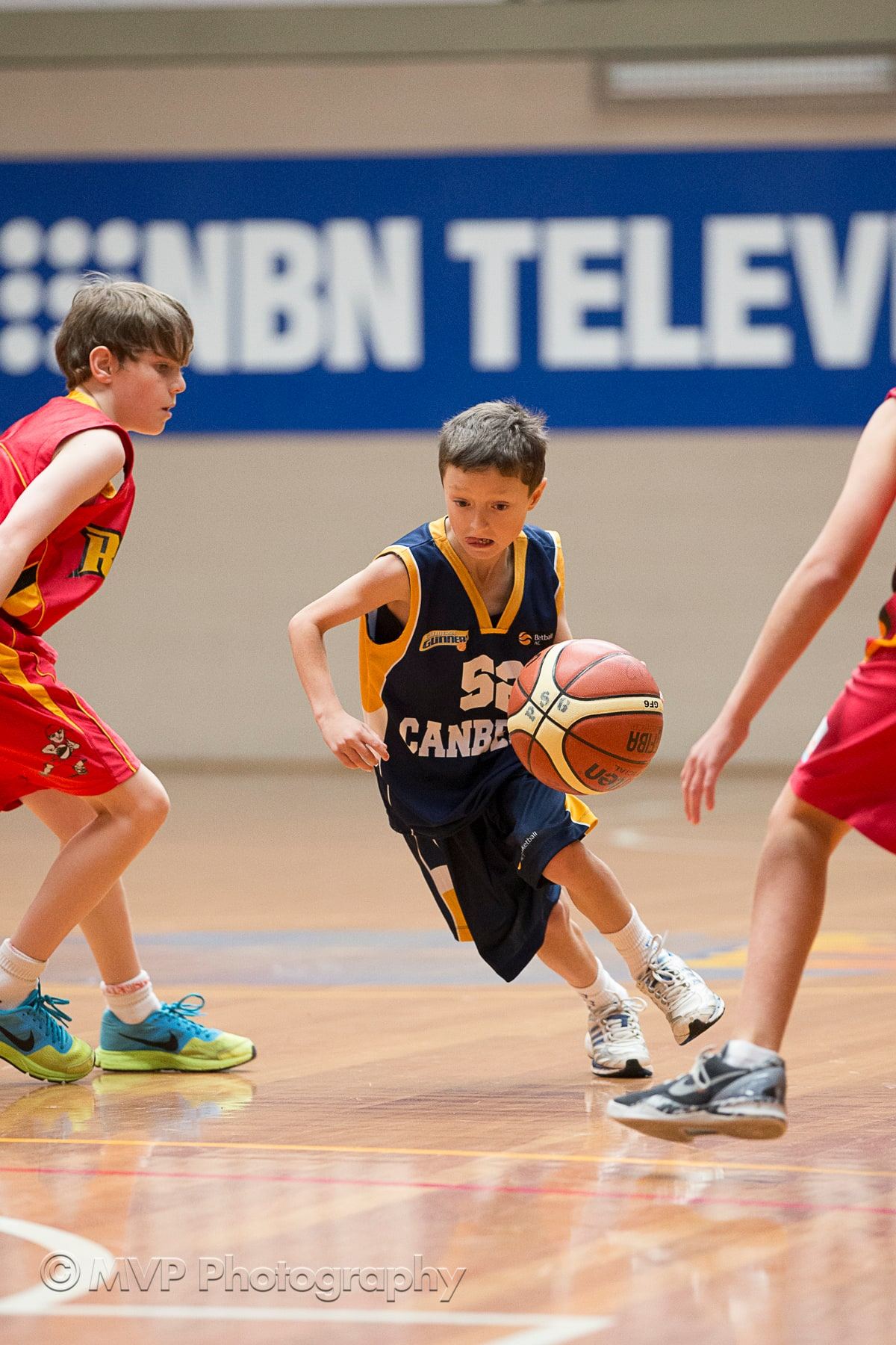 A young player in a Canberra Gunners jersey dribbles past two defenders in red on an indoor court.