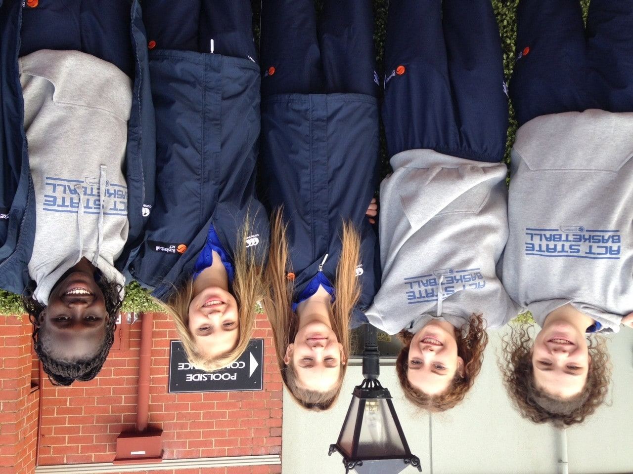 Five players in ACT State Basketball jackets and hoodies smile together outside a building.