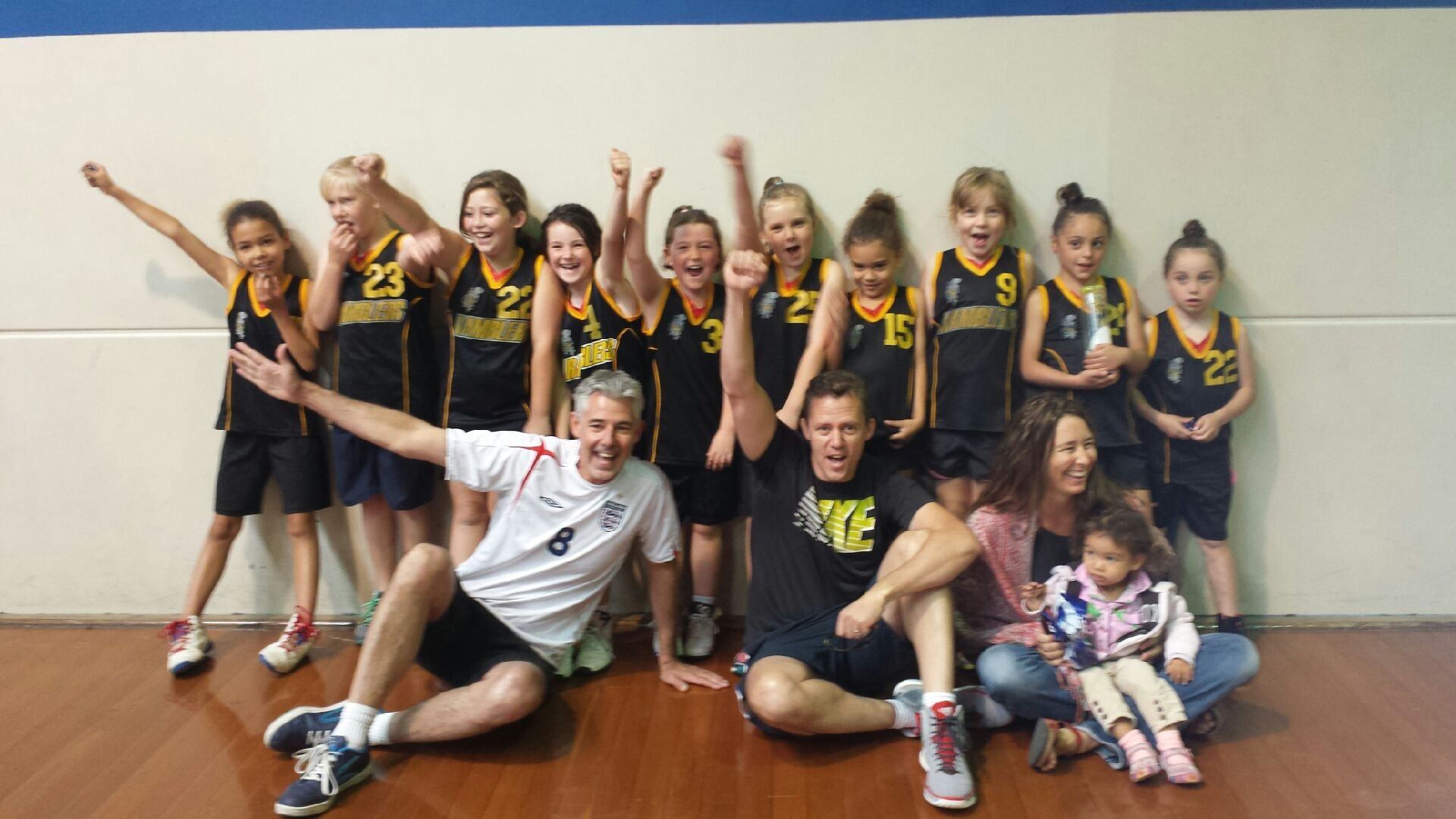 A Ramblers junior girls' team cheer with raised fists alongside coaches and family, celebrating a win.