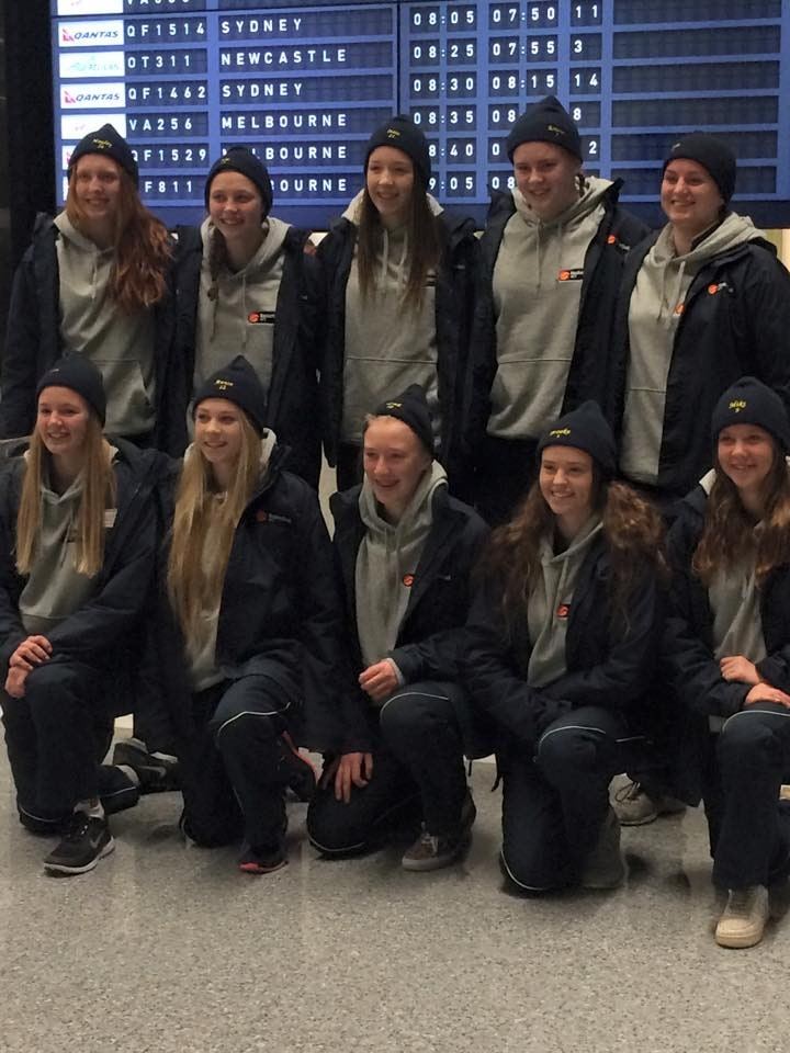 The ACT U16 girls' team pose together in team jackets and beanies at the airport, departures board behind them.