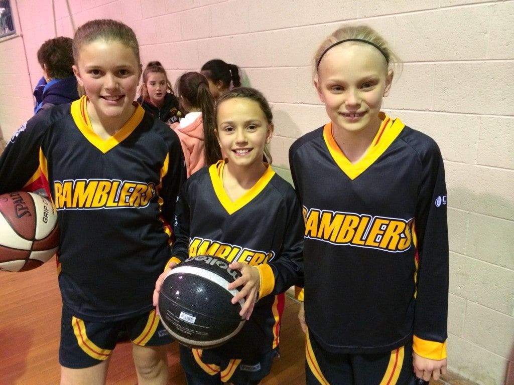 Three smiling Ramblers junior players in black and gold jerseys, two holding basketballs, stand together courtside.
