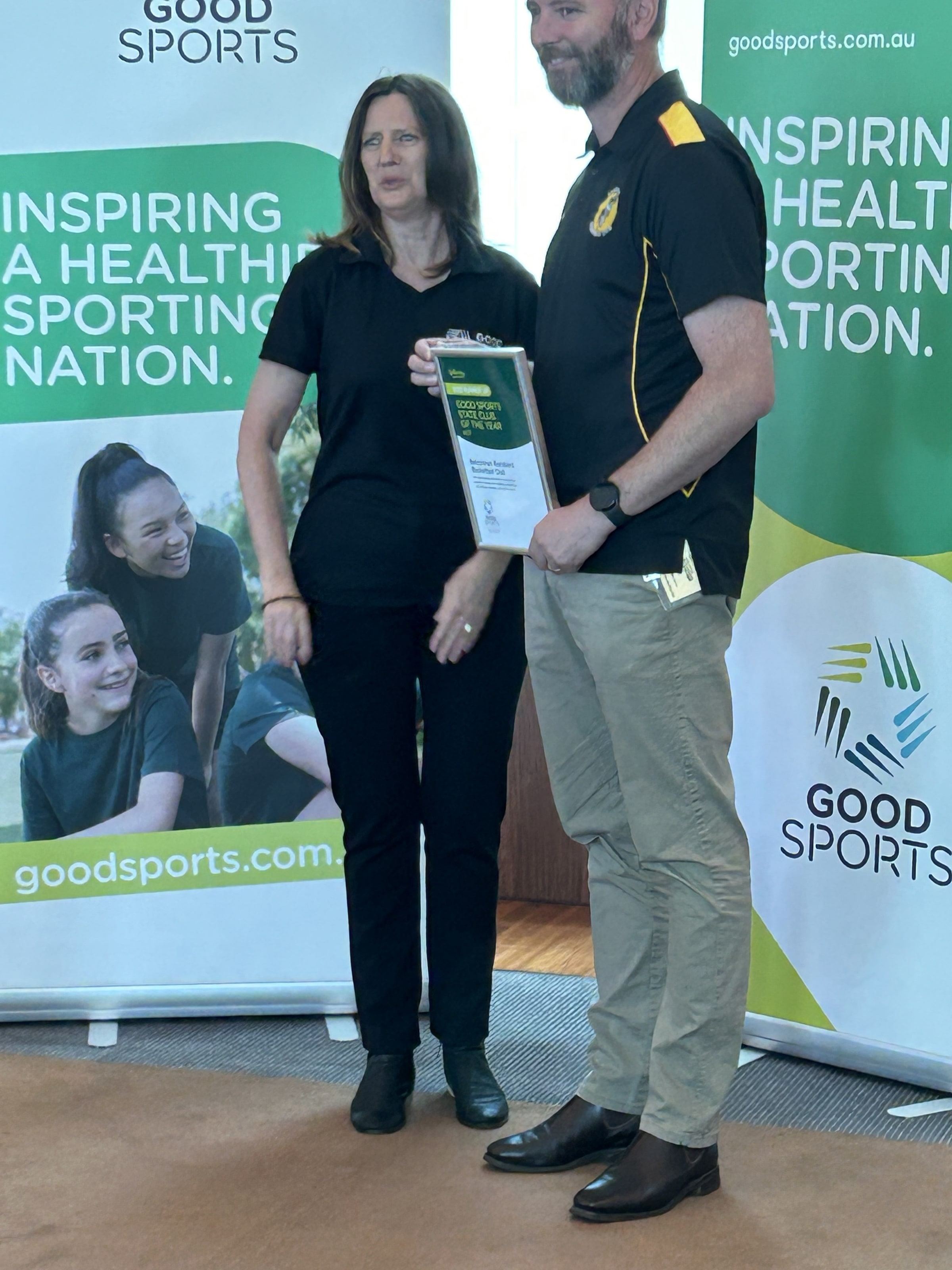 A club representative in a Ramblers polo accepts a framed Good Sports award certificate from a presenter, banners behind them.