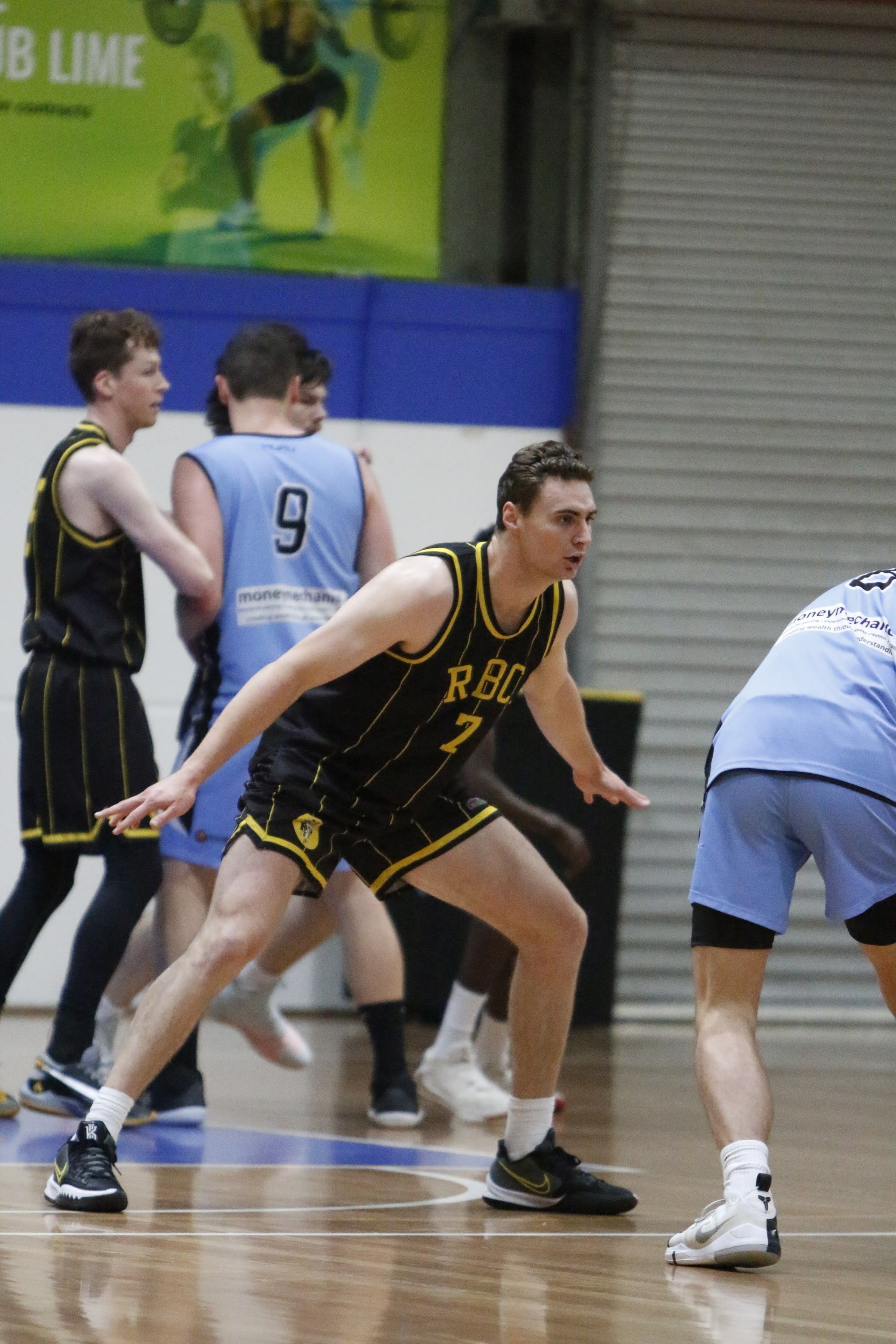 A Ramblers senior player in black and gold sets a defensive stance, opponents in light blue nearby.