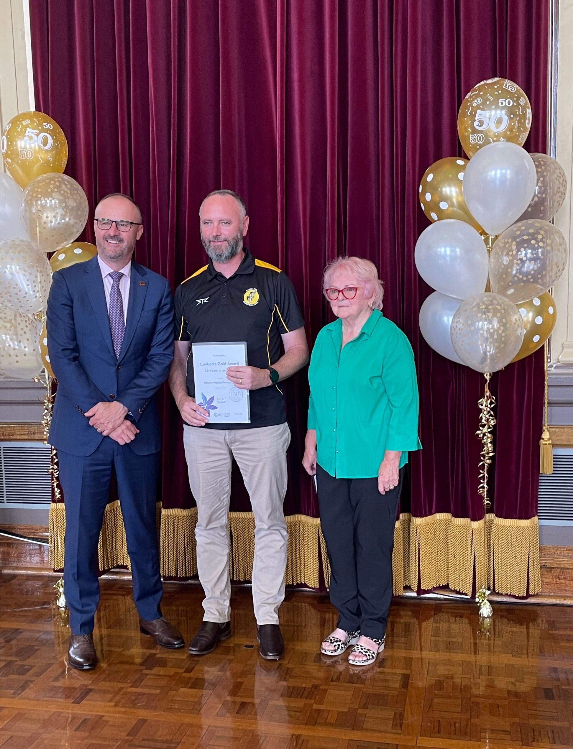 The club president holds the Canberra Gold Award certificate, flanked by a presenter and a club life member, gold balloons on stage behind them.