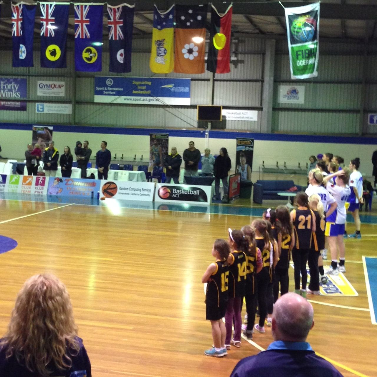 Junior girls in black and gold stand courtside under the venue's flags ahead of the national anthem, senior players and officials nearby.