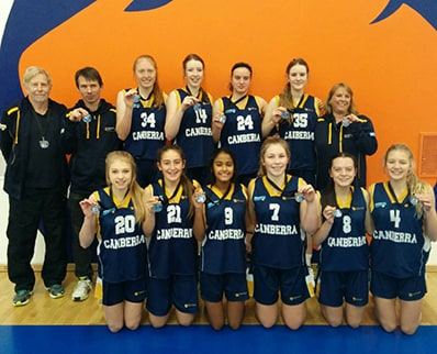 The ACT U16 girls' state basketball team pose together in navy Canberra uniforms wearing their selection medals, with two coaches.